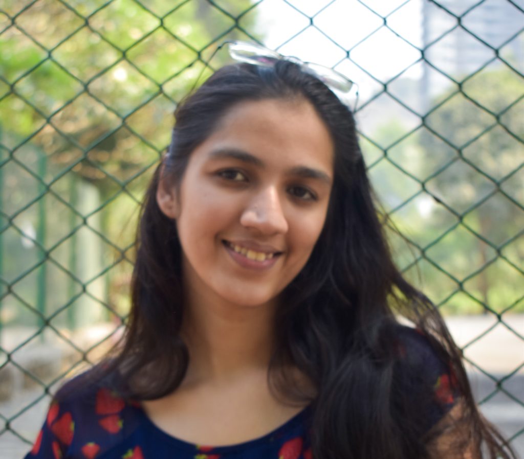 Monaami Pal smiles at the camera. She is wearing a blue shirt with strawberries. She is standing in front of a chain link fence and a park can be seen behind her.