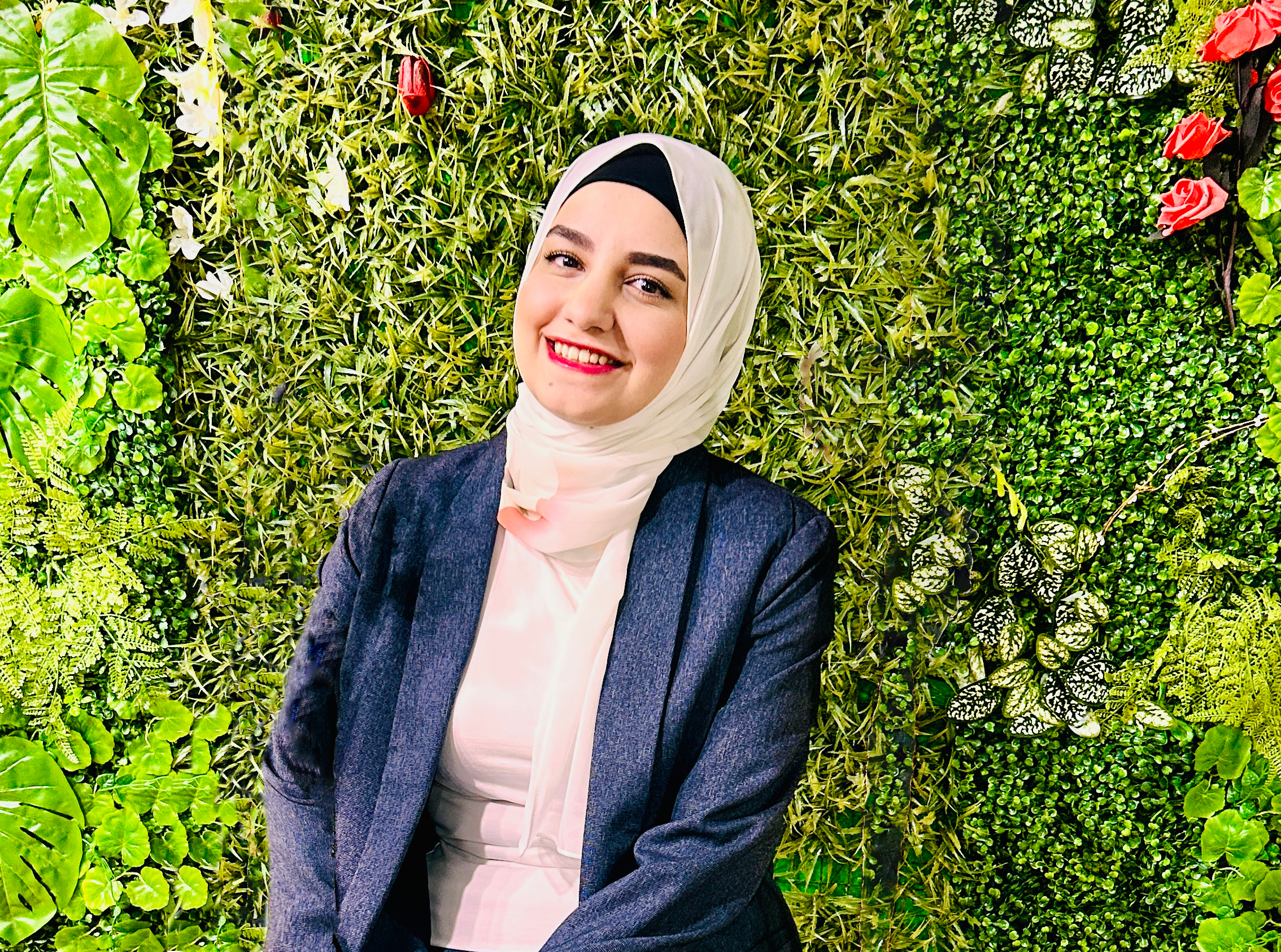Sondos smiles at the picture. She is sitting down, wearing a light beige hijab, light pink shirt, and dark blue blazer. The background is a multitude of artificial green plants, with some red roses in the top right.